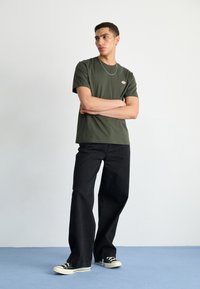 Tricou din bumbac de culoare verde-oliiv cu mâneci scurte și un mic logo, asortat cu blugi negri cu picioare largi și adidași negri și albi.