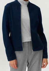 Persona che indossa una giacca zip blu navy sopra un dolcevita grigio chiaro e pantaloni grigi, in piedi davanti a uno sfondo semplice.