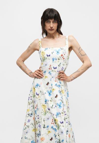 Γυναίκα με σκούρα μαλλιά και τατουάζ φοράει ένα λευκό καλοκαιρινό φόρεμα με μπλε, κίτρινα και κόκκινα λουλούδια, στέκεται με τα χέρια στα ισχία μπροστά σε λευκό φόντο.