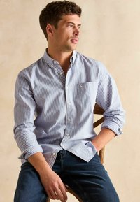 Koszula z długim rękawem zapinana na guziki w jasnoniebieskie i białe paski, z rękawami podwiniętymi do łokci, pojedynczą kieszonką na piersi oraz miękką bawełnianą tkaniną.