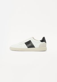 Sneaker bianco con accento in pelle nera, linguetta del tallone in suede beige testurizzato, design con lacci, suola in gomma e dettagli cuciti sui lati.