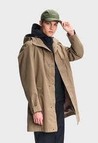 Homme ajustant une casquette vert olive, portant un imperméable à capuche beige par-dessus une veste noire à fermeture éclair et un pantalon foncé, sur un fond clair uni.