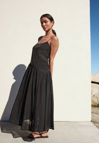 Robe maxi noire à fines bretelles, en tissu texturé, taille marquée et jupe fluide. Associée à des sandales plates noires.