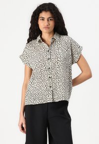 Camicia a maniche corte, con bottoni, di colore crema con motivo maculato nero. Tessuto morbido con vestibilità rilassata, dotata di due taschini sul petto.