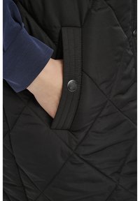 Veste noire matelassée avec des coutures diagonales, dotée d'une poche latérale avec un bouton pression argenté et une chemise à manches longues bleu marine qui dépasse.