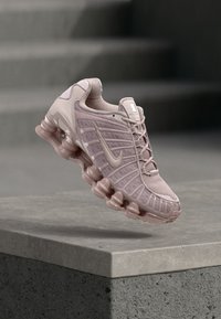 Αθλητικό παπούτσι Nike σε ανοιχτό ροζ ύφασμα με υφές επικάλυψης, ανακλαστικές λεπτομέρειες και σχεδιασμό σόλας με φυσαλίδες, κρεμασμένο πάνω από γκρί επιφάνεια.