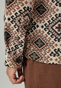 Camicia tessuta color crema con motivi geometrici in nero, rosso e marrone. Dettaglio con polsini abbottonati e tessuto testurizzato. Pantaloni marroni visibili.