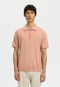 Jeune homme portant un polo zippé à manches courtes de couleur rose clair et un pantalon beige, debout devant un fond blanc uni.