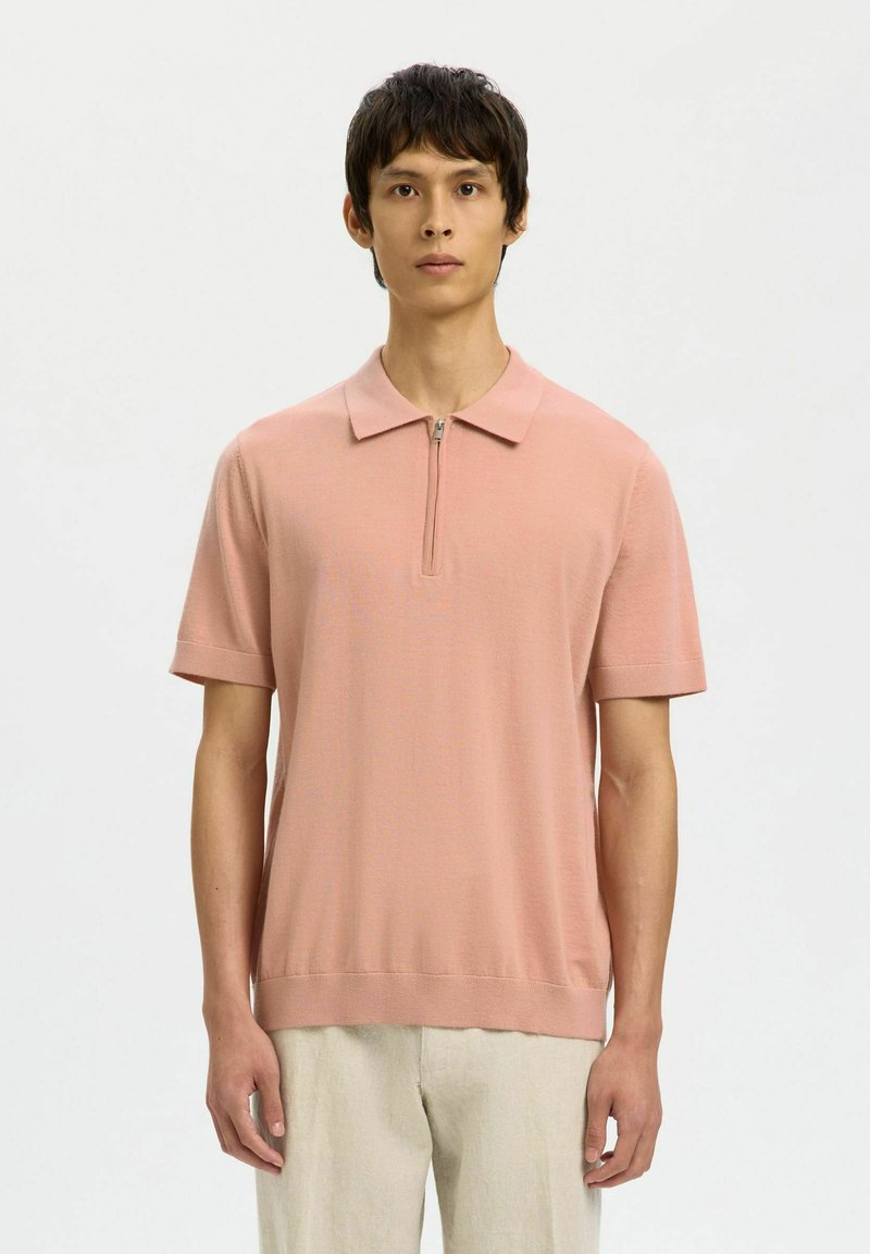 Jeune homme portant un polo zippé à manches courtes de couleur rose clair et un pantalon beige, debout devant un fond blanc uni.