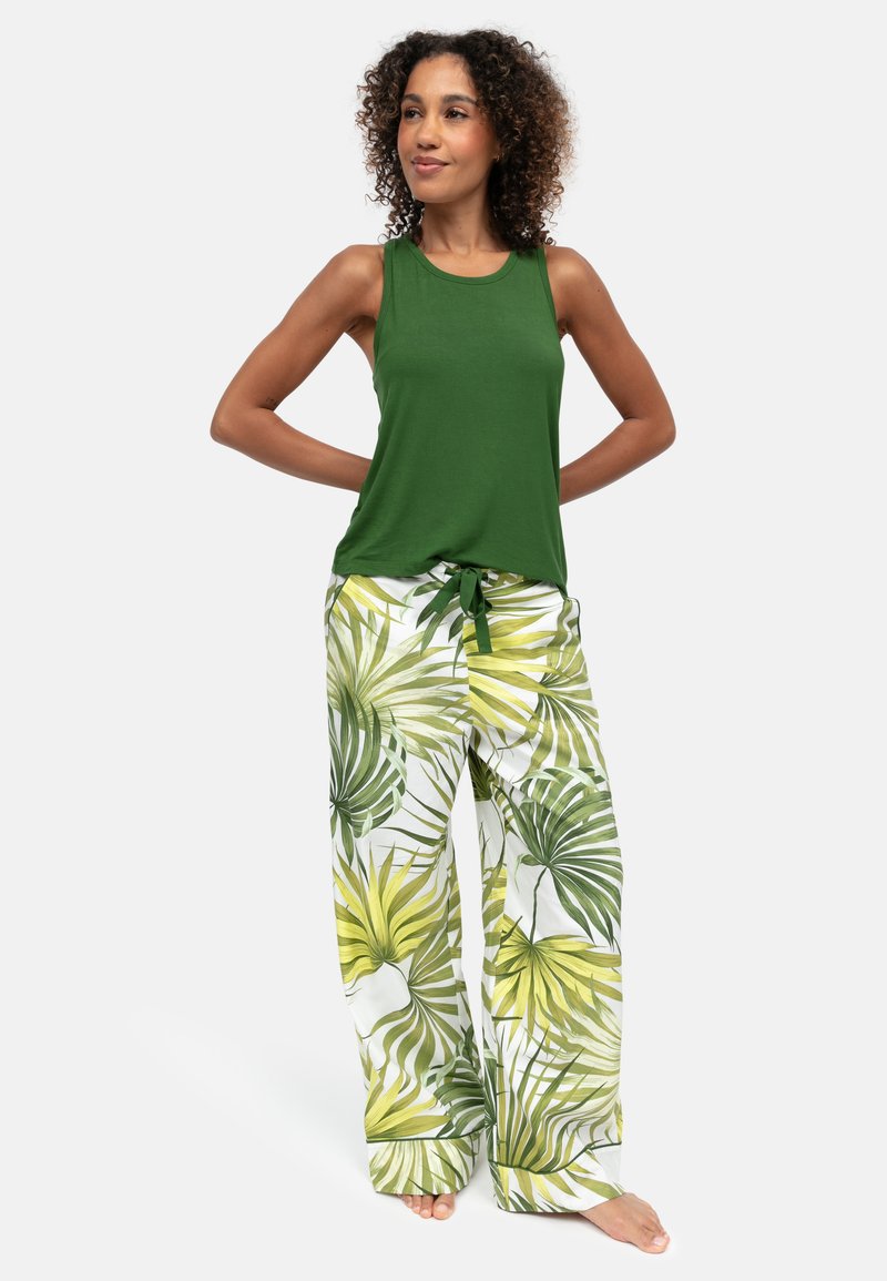 Frau steht barfuß, trägt ein ärmelloses grünes Oberteil und weiße, weit geschnittene Hosen mit grünen und gelben tropischen Blattmustern.