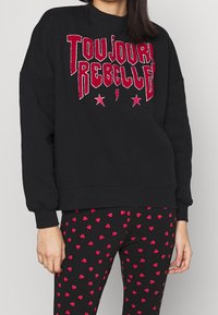 Sudadera negra con texto brillante rojo que dice "TOUJOURS REBELLE" y pequeños acentos de estrella y rayo, combinada con leggings negros con corazones rojos.