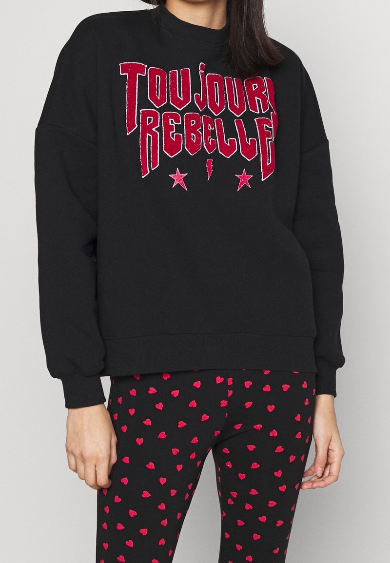 Sudadera negra con texto brillante rojo que dice "TOUJOURS REBELLE" y pequeños acentos de estrella y rayo, combinada con leggings negros con corazones rojos.