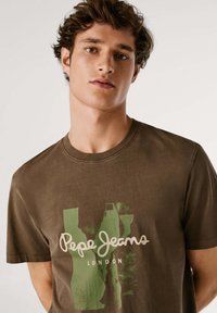 Camiseta de algodón marrón con logo blanco de "Pepe Jeans" y estampado gráfico verde. Cuello redondo, mangas cortas, textura suave.