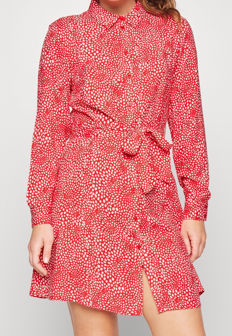 Robe chemise rouge à manches longues avec motif de points blancs irréguliers, boutonnée sur le devant, ceinture nouée à la taille, portée par une personne debout.