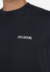 Zwarte katoenen t-shirt met ronde hals en korte mouwen, met een wit "PEGADOR" logo geborduurd op de linkerborst.