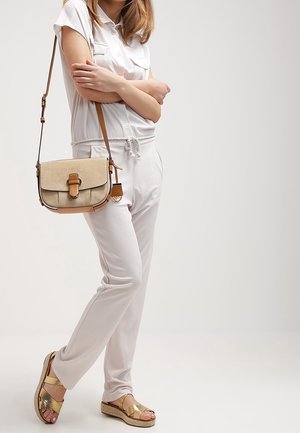Witte ontspannen jumpsuit met korte mouwen, een trekkoord in de taille en een lichtbruine crossbody tas. Gouden accenten op espadrille sandalen.