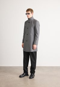 Pelēks vilnas mētelis ar augstu apkakli un pogu aizdari, apvienots ar melniem biksēm un spīdīgām melnām kurpēm. Modelis valkā tumšas saulesbrilles.