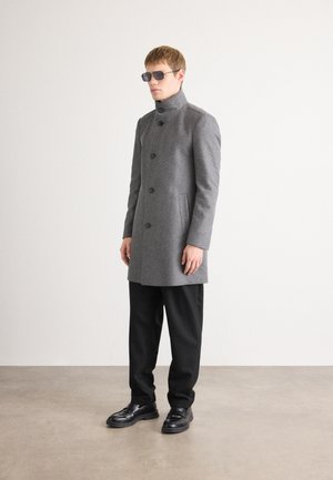 Manteau en laine gris avec un col montant et une fermeture à boutons, associé à un pantalon noir et des chaussures noires brillantes. Le modèle porte des lunettes de soleil noires.