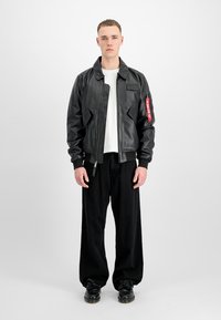 Jeune homme debout portant une veste en cuir noire avec une étiquette rouge sur le bras, un t-shirt blanc, un pantalon noir large et des chaussures noires sur fond blanc.