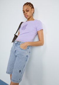 Levendula crop póló grafikai tervezéssel, világoskék farmer bike shorts-szal párosítva, amelynek az alján kis hímzett részlet található.