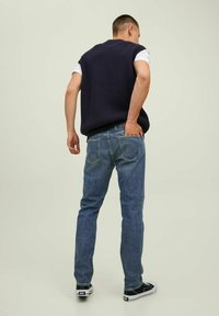 Maglione senza maniche a coste in marina, T-shirt bianca sotto, jeans in denim blu con tasche posteriori e sneakers nere. Sfondo chiaro e semplice.