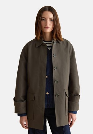 Mujer con cabello castaño liso lleva un abrigo de botones sobredimensionado color verde oliva sobre denim azul y camisa a rayas, de pie frente a fondo blanco.