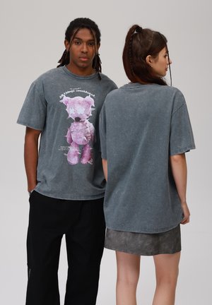 Gray oversized T-shirt featuring a graphic of a pink teddy bear with stitched accents, worn by models facing both directions.