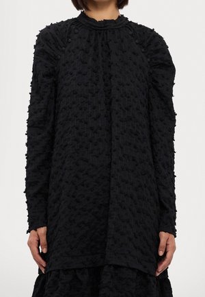 Mujer con vestido negro texturizado de manga larga, con cuello alto y patrón de puntos elevados.