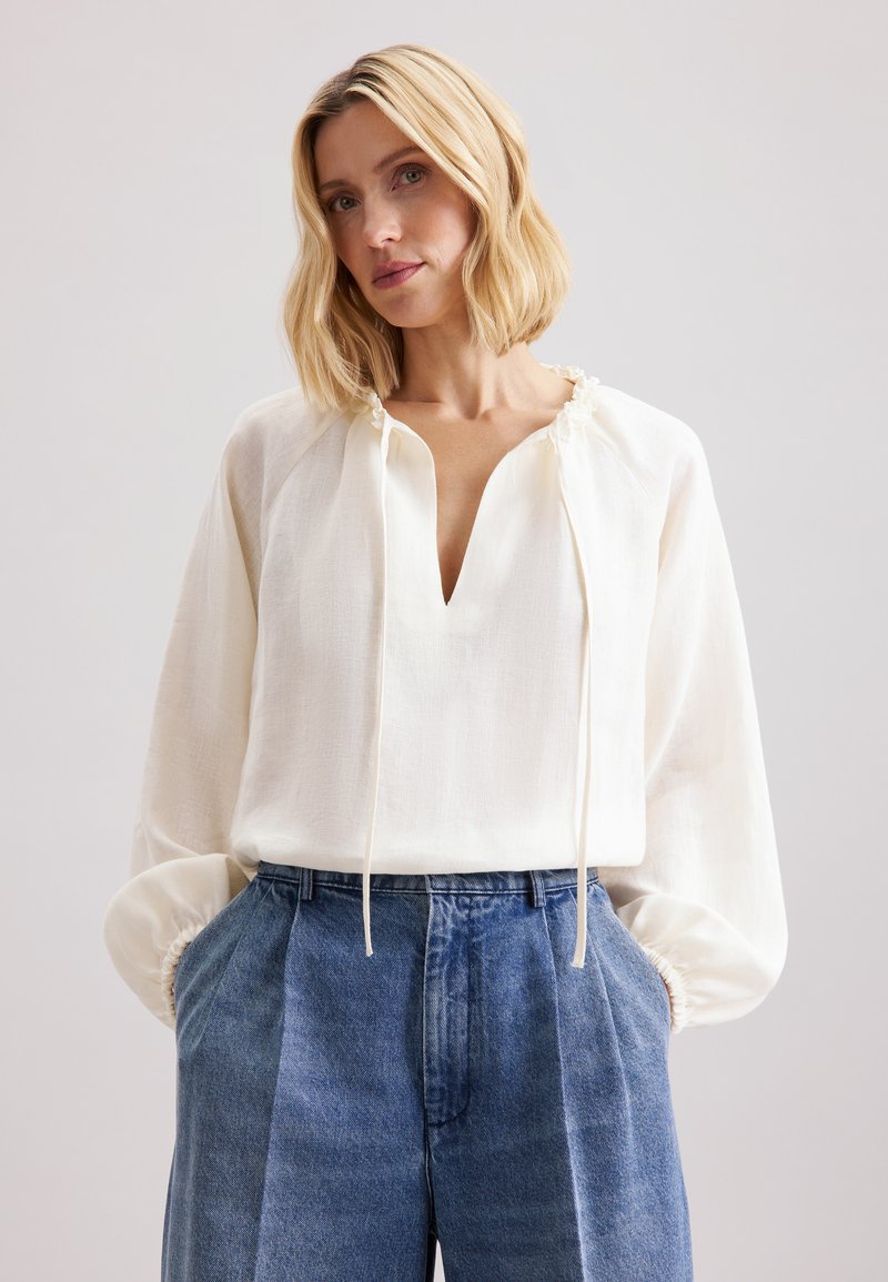 Blusa bianca a maniche lunghe con scollo a V e colletto con lacci; tessuto morbido e leggero e polsini elasticizzati. Indossata con pantaloni in denim blu a vita alta.
