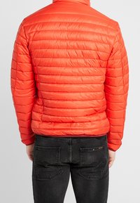 Doudoune rouge rembourrée avec des lignes de matelassage horizontal, en matériau léger, col montant et poignets élastiques, portée sur un jean en denim foncé.