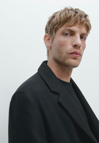 Cappotto nero con un colletto strutturato, tessuto testurizzato e una vestibilità rilassata. Il modello ha capelli biondi corti e testurizzati e una camicia neutra sotto.