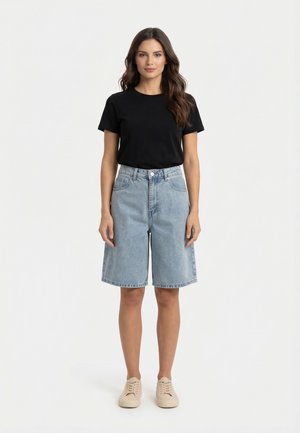 Donna in piedi di fronte, indossa una maglietta nera a maniche corte, pantaloncini di jeans azzurri fino al ginocchio e scarpe da ginnastica beige su sfondo bianco.