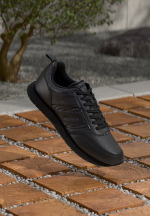 Svarte Calvin Klein lær sneakers med lisser, vist over trefliser og små grus utendørs nær et tre og betongdekke.