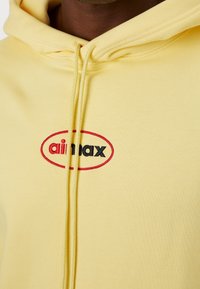 Gros plan d’une personne portant un sweat à capuche jaune avec un logo "air max" rouge et noir brodé sur la poitrine.