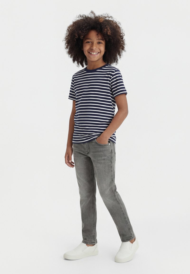 Dítě s kudrnatými vlasy se usmívá, nosí tmavě modré a bílé pruhované tričko, šedé džíny a bílé nazouvací boty, stojí s jednou rukou v kapse.