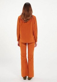 Ensemble en velours côtelé rouille en deux pièces avec une chemise à manches longues boutonnée et un pantalon évasé. Texture lisse et couleur orange chaud.