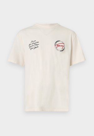 Bež bombažna majica z okroglim izrezom, kratkimi rokavi in vezenimi logotipi. Besedilo v črni in rdeči barvi vključuje imena mest in blagovno znamko.