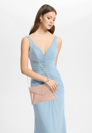 Jeune femme en robe bleu clair plissée sans manches, tenant une pochette beige à chaîne, sur fond blanc.