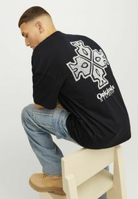 Crna pamučna majica s dekorativnim bijelim geometrijskim uzorkom na leđima, koja sadrži tekst "Originals Studio" na dnu.