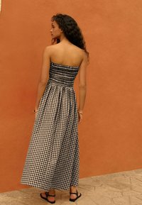 Czarno-biała sukienka maxi w kratkę gingham z bezrękawą, elastyczną górą i zwiewnym dołem, zestawiona z czarnymi sandałami na tle pomarańczowej ściany.