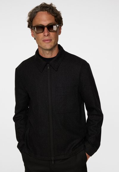 Svart zip-jacka med en strukturerad krage, en bröstficka och texturerad tyg, matchad med rektangulära solglasögon.