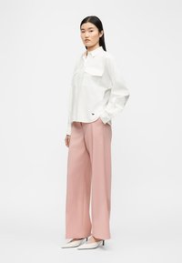 Mujer de pie con una camisa blanca de manga larga con botones, pantalones anchos rosa y tacones puntiagudos blancos sobre un fondo blanco liso.