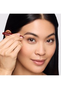 Pince à épiler rouge avec une pointe effilée, tenue par une main près d'un sourcil, sur un fond neutre. Texture lisse, design ergonomique.