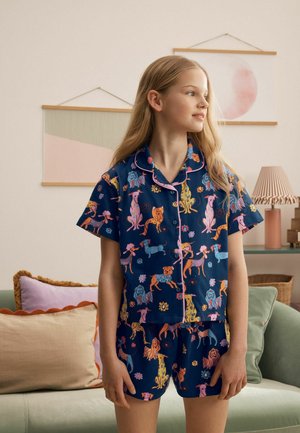 Pige iført marineblåt pyjamassæt med korte ærmer og farverige hundetryk, stående indendørs nær en sofa med puder og indrammet vægkunst.