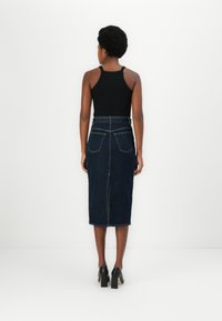Jupe en denim avec une taille ajustée, de longueur intermédiaire, couleur bleu foncé, cinq poches et fente arrière. Associée à un top sans manches noir et des talons.