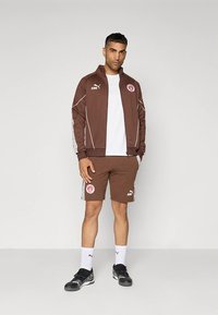Ensemble de sport marron avec accents et logo blancs, comprenant une veste zippée et un short, associé à des chaussures noires et des chaussettes blanches.