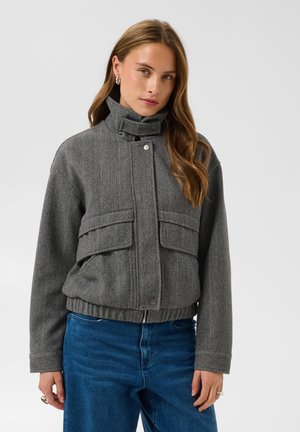 Femme aux cheveux longs portant une veste gris chevron à col montant et un jean bleu, debout devant un fond clair uni.