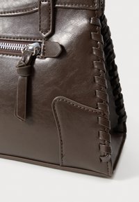 Hnedá kožená kabelka so štruktúrovaným povrchom, vybavená zipsom, zapletenými ozdobami a pevnou konštrukciou. Na boku je viditeľná detailná kapsa.