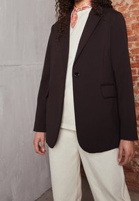 Blazer marrone su misura con rever e chiusura a un bottone. Indossato sopra una camicia bianca e abbinato a pantaloni chiari.