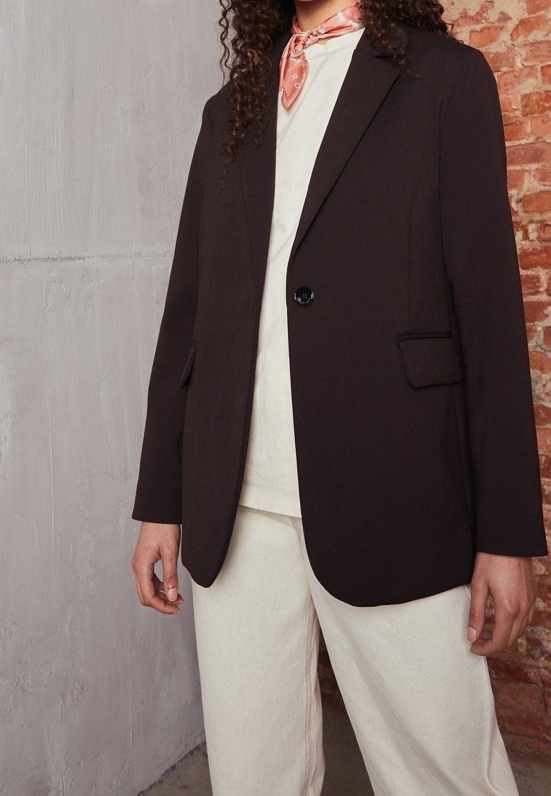 Blazer marrone su misura con rever e chiusura a un bottone. Indossato sopra una camicia bianca e abbinato a pantaloni chiari.